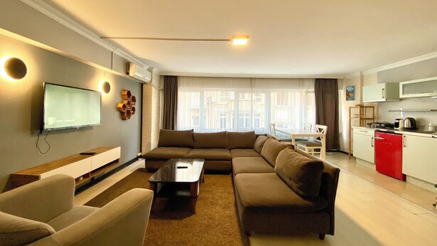 A modern living room with a sectional sofa, a flat-screen TV, and a kitchen area with a red refrigerator. - M4 Istiklal Spacious and Central 1+1 Apartment (Istanbul)