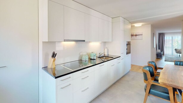 Una cocina moderna con armarios blancos, encimera negra y un comedor con mesa de madera y sillas azules.