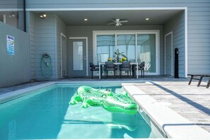 Una piscina con un flotador de cocodrilo inflable verde, un área de patio con mesa y sillas, y una zona de estar cubierta con ventilador de techo.