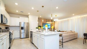 A modern kitchen with white cabinetry, stainless steel appliances, a large island, and a seating area with a sofa and chairs. - Family 5Bd Close To Disney At Windsor Cay 16136 (Clermont)