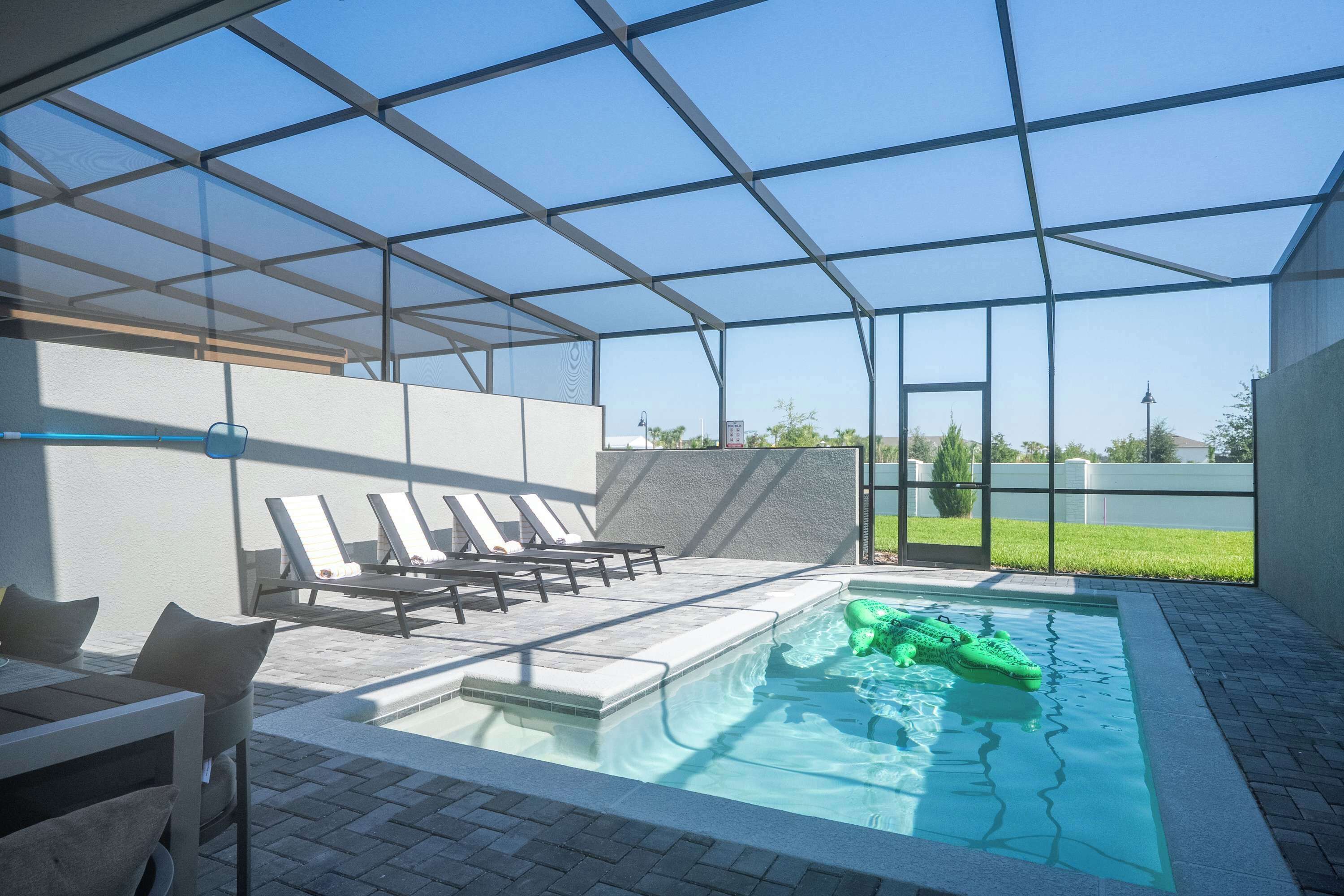 Zona de piscina con sillas de descanso, una estructura de vidrio y un cocodrilo hinchable verde.