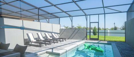 Zona de piscina con sillas de descanso, una estructura de vidrio y un cocodrilo hinchable verde.