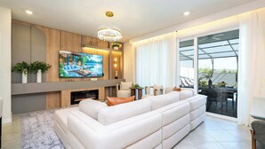 A modern living room with a large TV, a fireplace, a sofa, and a dining area with a glass door leading outside. - Family 5Bd Close To Disney At Windsor Cay 16136 (Clermont)