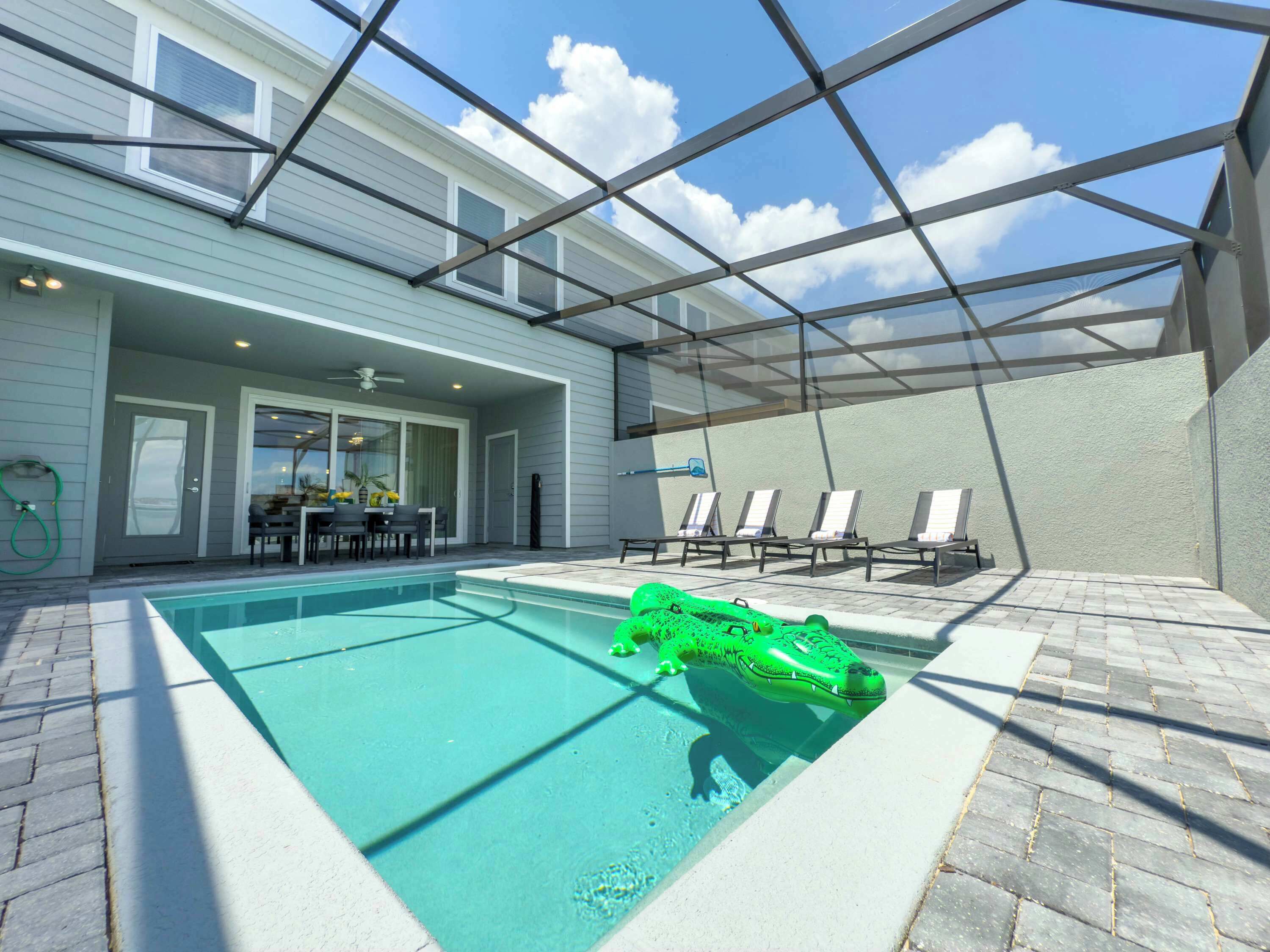 A swimming pool with a green inflatable crocodile toy, a covered patio area with chairs, and a view of the sky through a metal roof structure.