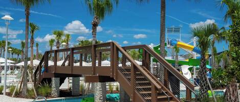 A pool area with a wooden bridge, palm trees, and a water slide.