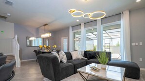 A modern living room with a sectional sofa, a glass coffee table, and a large window with curtains. - Magical 5Br W Pool Minutes From Disney 16140 (Clermont)