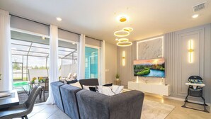 A modern living room with a grey sectional sofa, a dining table with chairs, a TV mounted on the wall, and a small table with a chair. - Magical 5Br W Pool Minutes From Disney 16140 (Clermont)