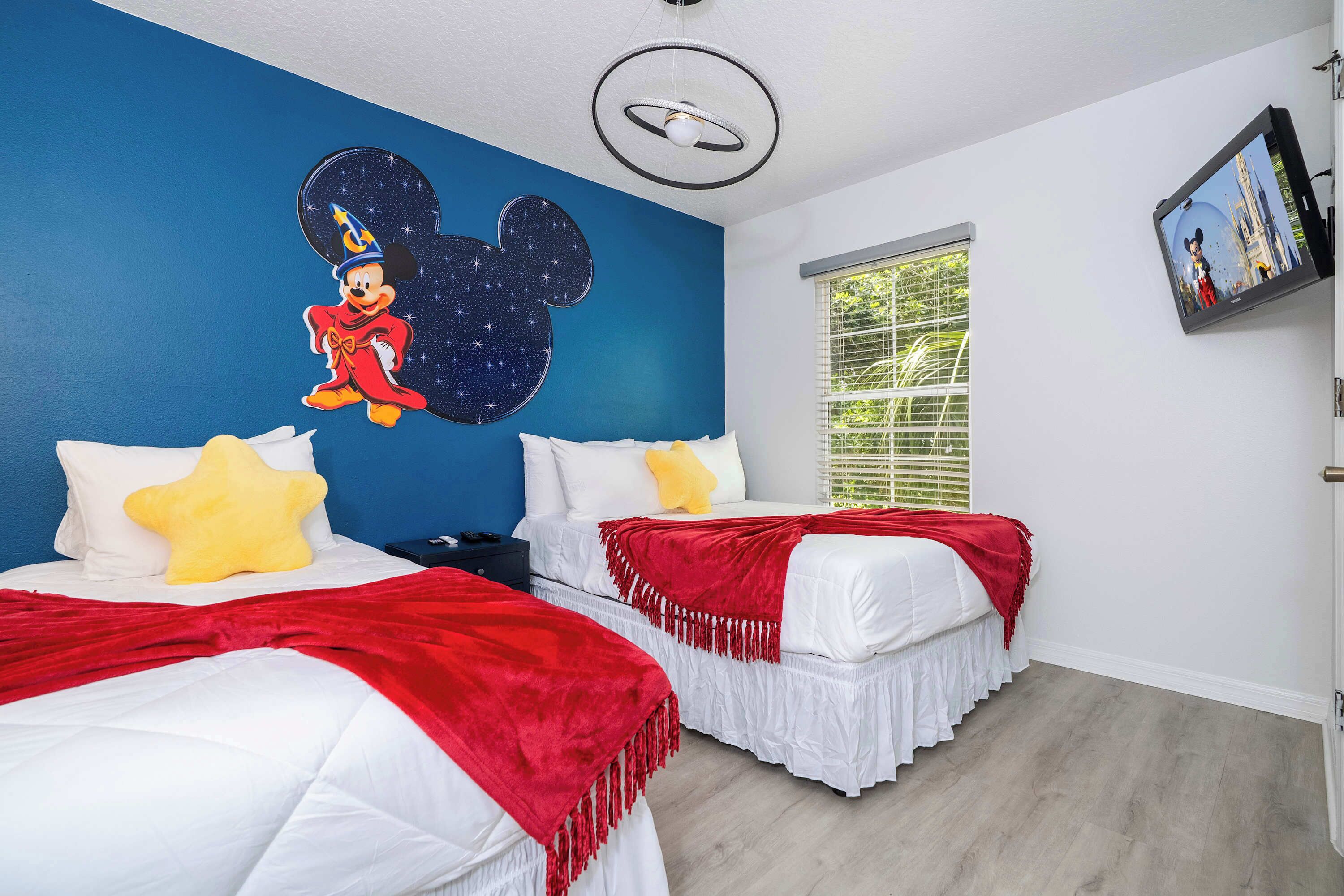 Habitación de hotel con dos camas, cada una con ropa de cama blanca y detalles rojos, un gran mural de Mickey Mouse en la pared y una ventana con persianas.