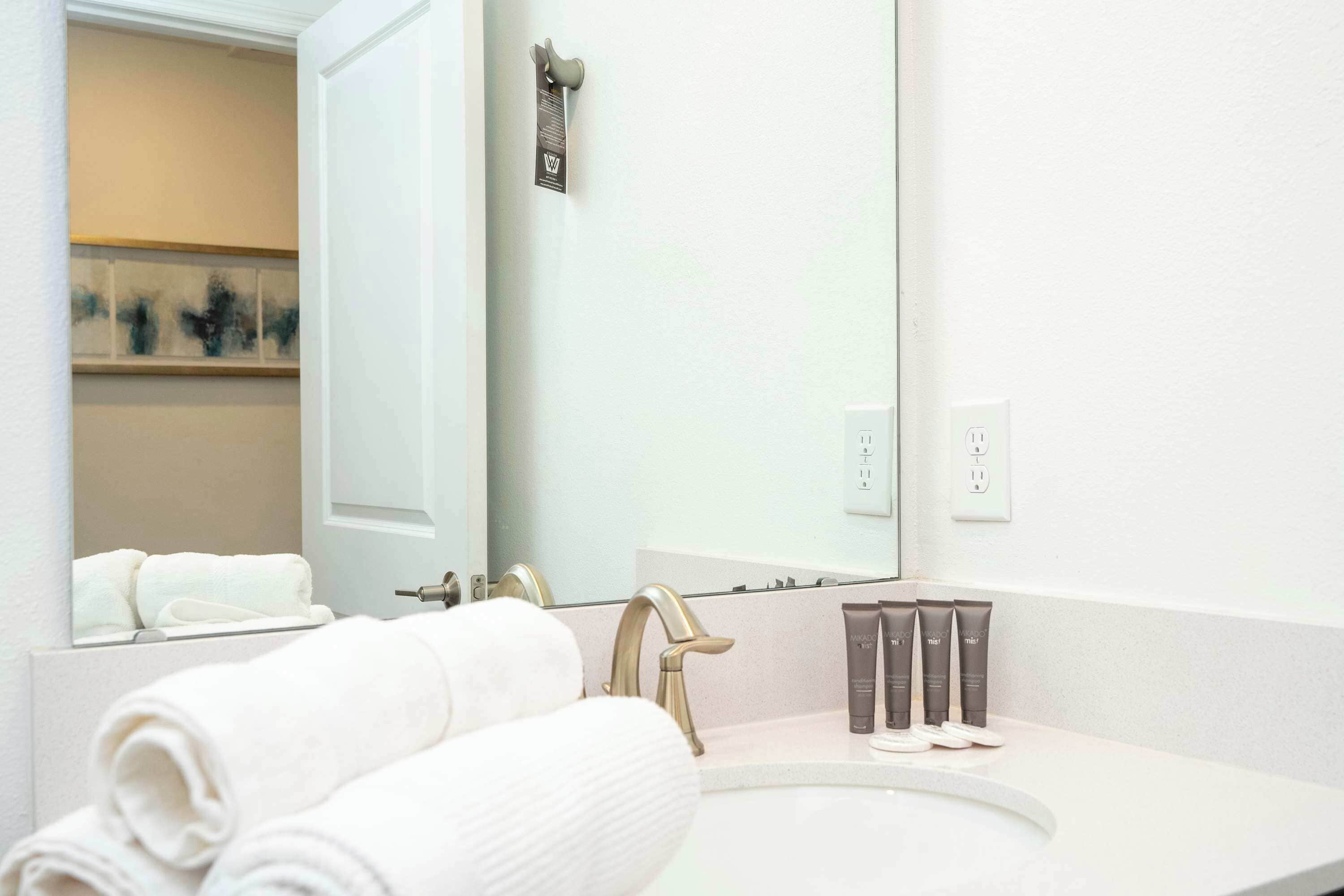 Un baño moderno con un lavabo blanco, grifo dorado y un espejo grande. Hay toallas y artículos de aseo en la encimera.