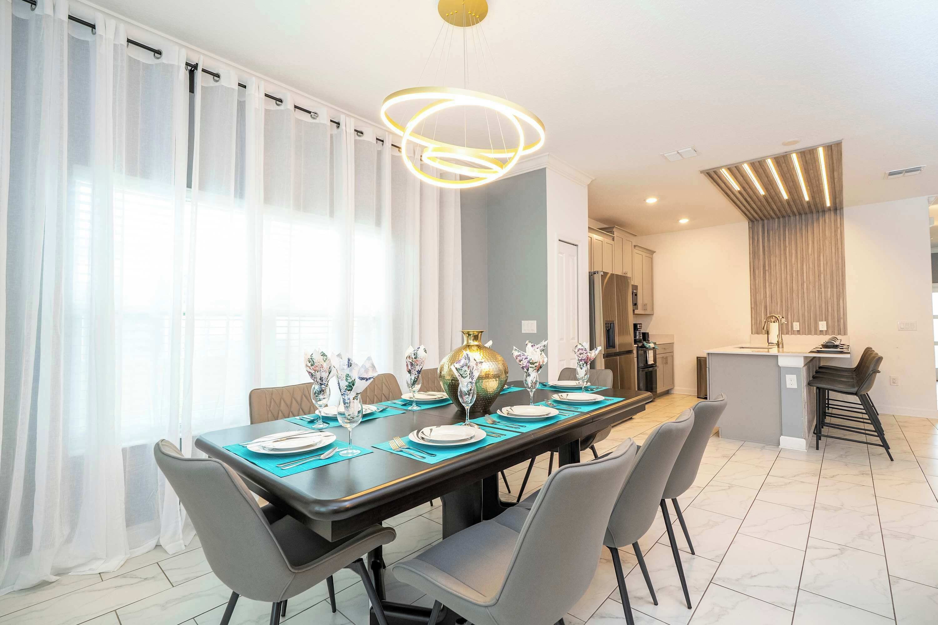 Een moderne eethoek met een lange tafel voor zes personen, een kroonluchter en een keuken op de achtergrond.