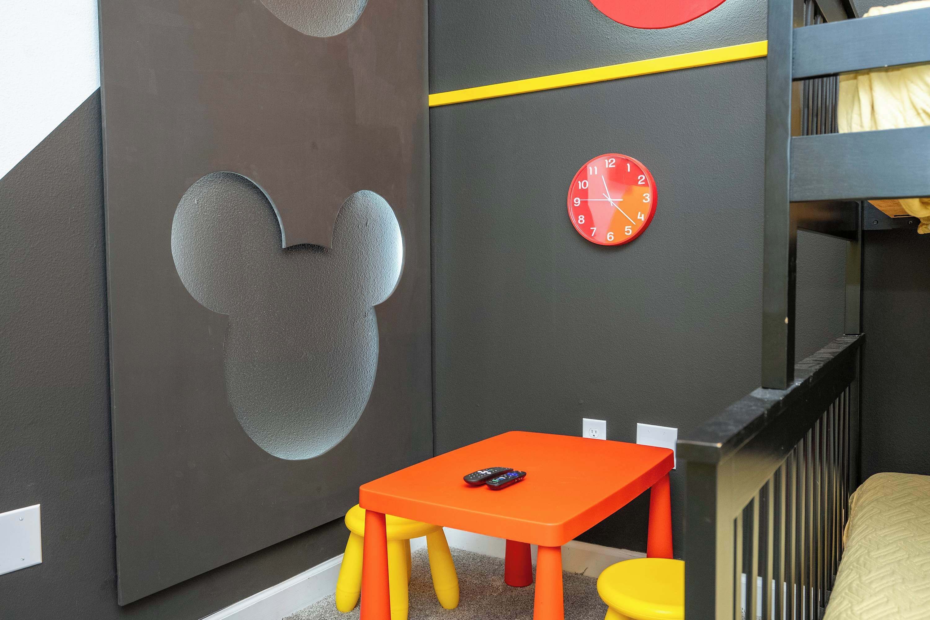 A child's room with a Mickey Mouse cutout, a red clock, and a small orange table with yellow legs.