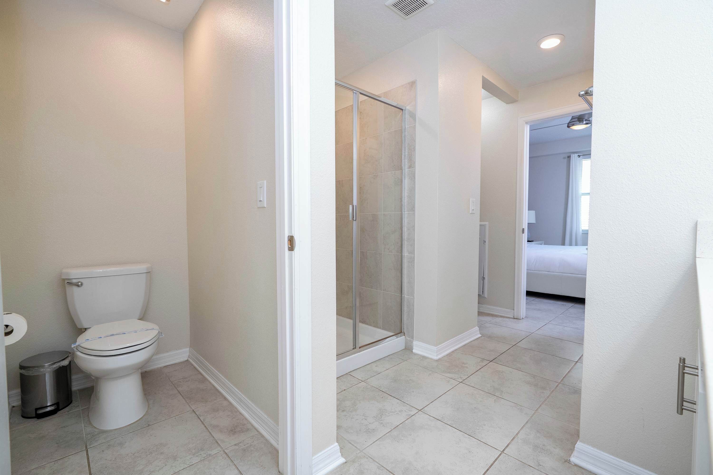 Une salle de bain avec des toilettes, une poubelle, une douche avec des portes en verre et un couloir menant à une chambre.