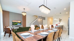 A modern dining area with a wooden table set for six, surrounded by upholstered chairs. A chandelier hangs above, and a flat-screen TV is mounted on the wall. - Vibrant 5Br W/ Pool & Waterpark @Storey Lake 4811M (Kissimmee)