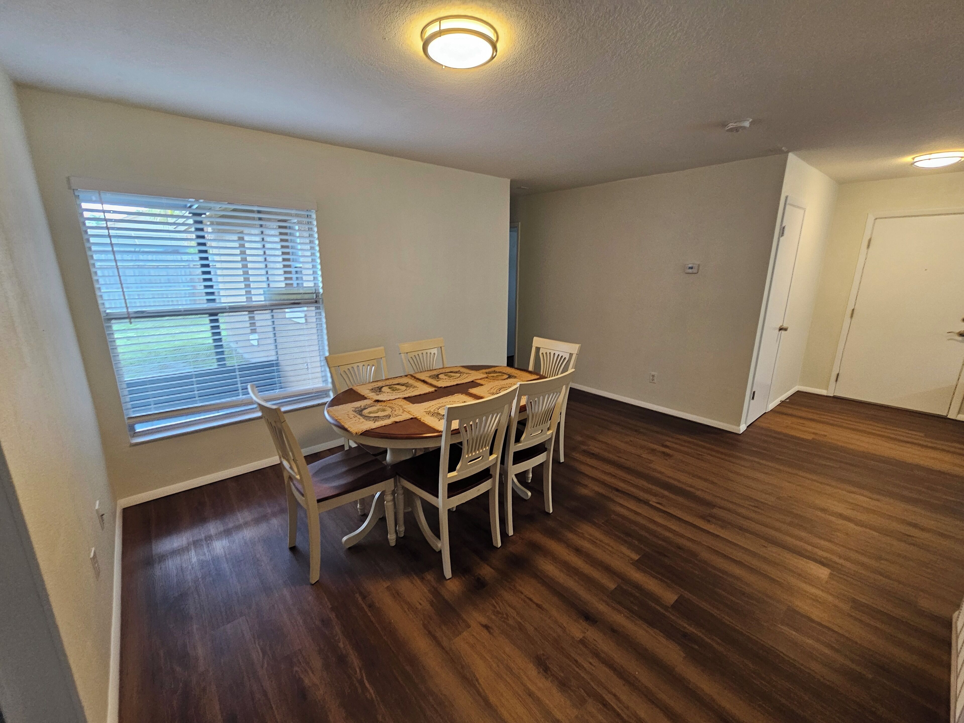 A dining area with a round table and six chairs, a window with blinds, and a door.