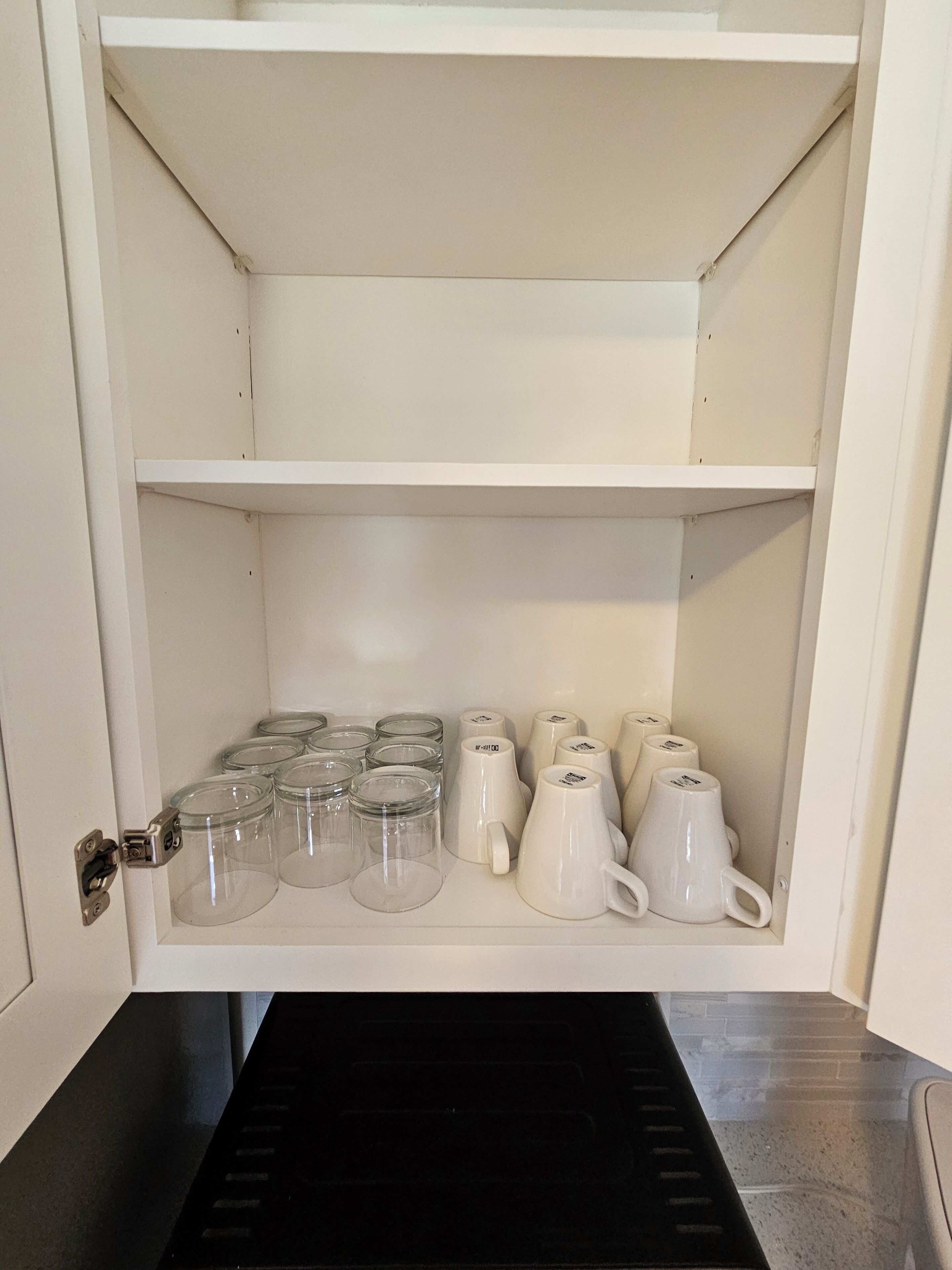 Un armadietto bianco con ripiani che contengono barattoli di vetro trasparenti e tazze di ceramica bianca.