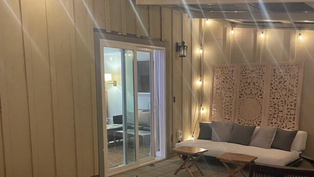 Ein überdachter Patio-Bereich mit einem Holztisch, einer Couch und einem Sessel. Lichterketten und eine dekorative Wandverkleidung sind vorhanden.