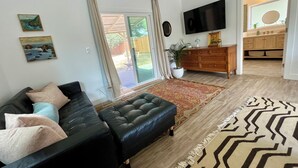 Ein Wohnzimmer mit einer schwarzen Ledersofa, einem gemusterten Teppich, einem Flachbildfernseher und einer Schiebetür zu einer Terrasse.