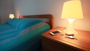 A neatly made bed with a striped blue and green bedspread, a bedside lamp, a smartphone, and a set of keys on a wooden nightstand. - Seaside Apartment in Rosolina Mare (Rosolina)
