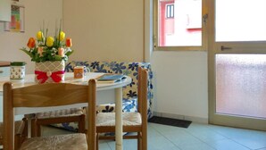 A dining area with a table set for two, a floral-patterned sofa, and a window with a view of a red building. - Seaside Apartment in Rosolina Mare (Rosolina)