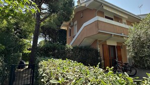 Ein zweistöckiges Gebäude mit Balkon, umgeben von Grünpflanzen und einem Tor.