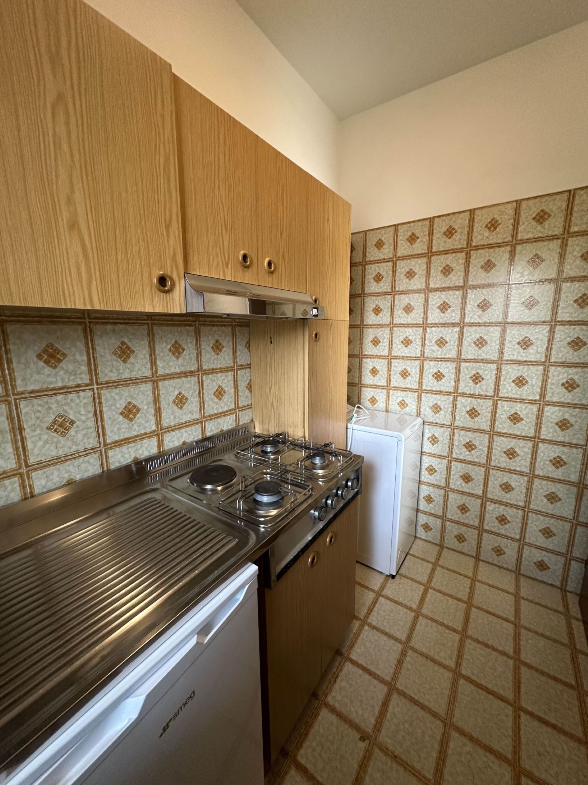 A compact kitchen with a gas stove, a built-in oven, and a white refrigerator. The kitchen has wooden cabinets and patterned tile backsplash.