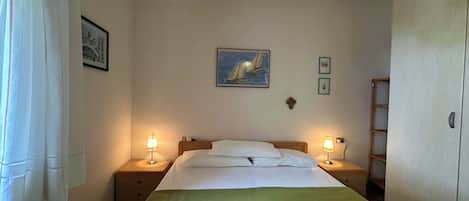 A bedroom with a bed, two bedside lamps, a painting, and a cross on the wall.