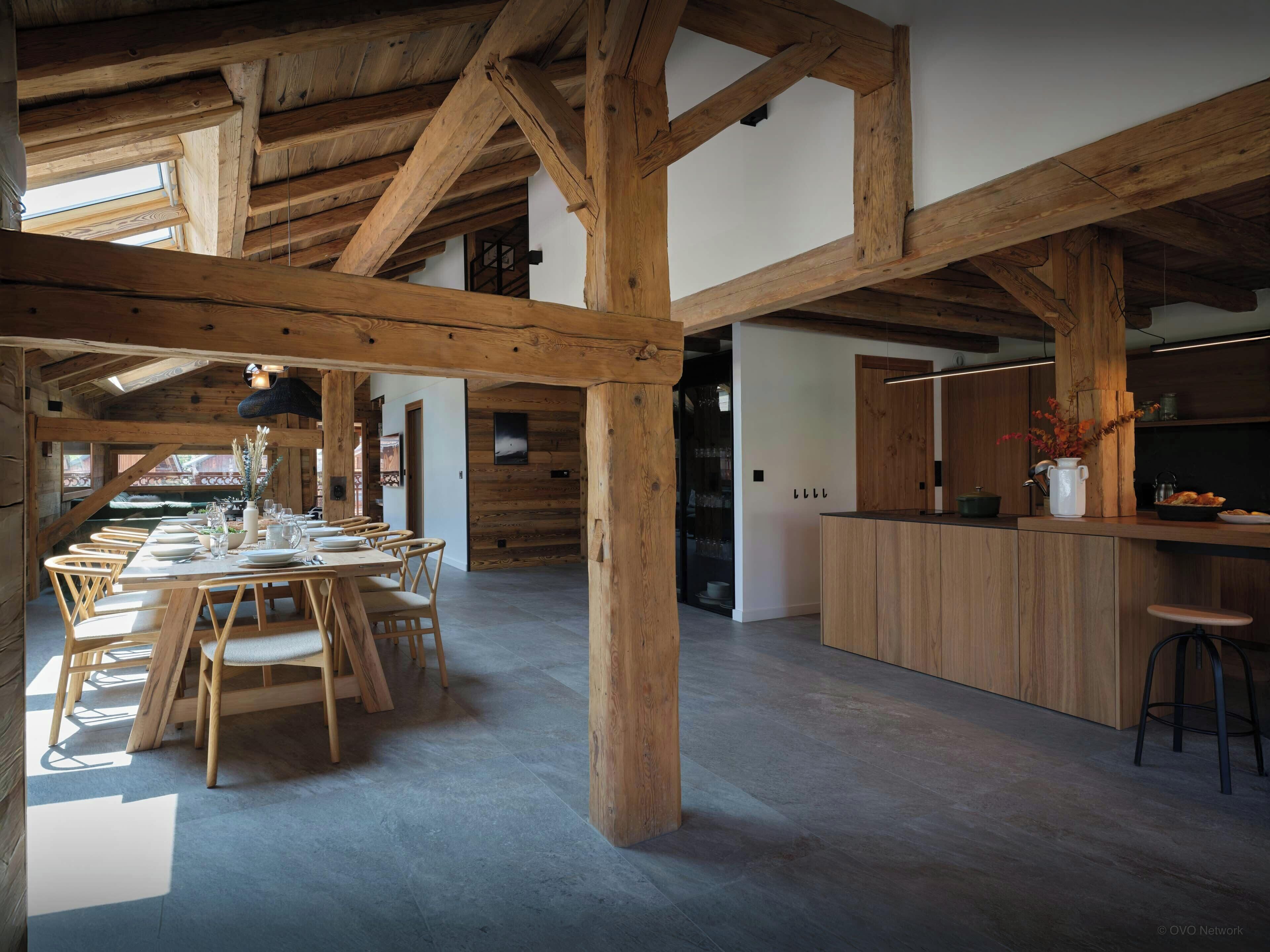 Une salle à manger spacieuse avec des poutres en bois, une longue table dressée pour accueillir plusieurs personnes, et un coin cuisine doté de meubles en bois et d’un plan de travail.