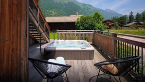 A wooden deck with a hot tub, two black chairs, and a view of mountains.