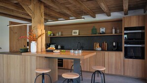 A modern kitchen with wooden beams, a central island with stools, built-in appliances, and a dark backsplash.