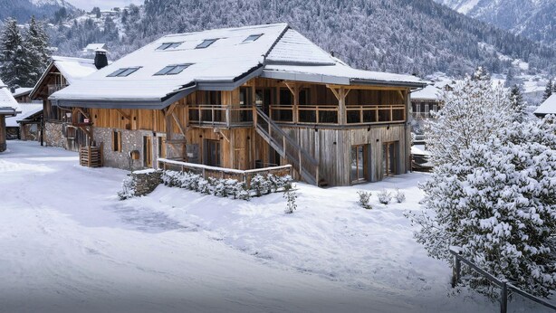 A wooden chalet with a snow-covered roof, a balcony, and a stone foundation.