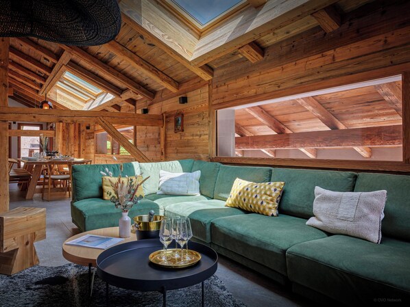 Un acogedor interior de cabaña de madera con un sofá verde, vigas de madera y ventanas amplias.