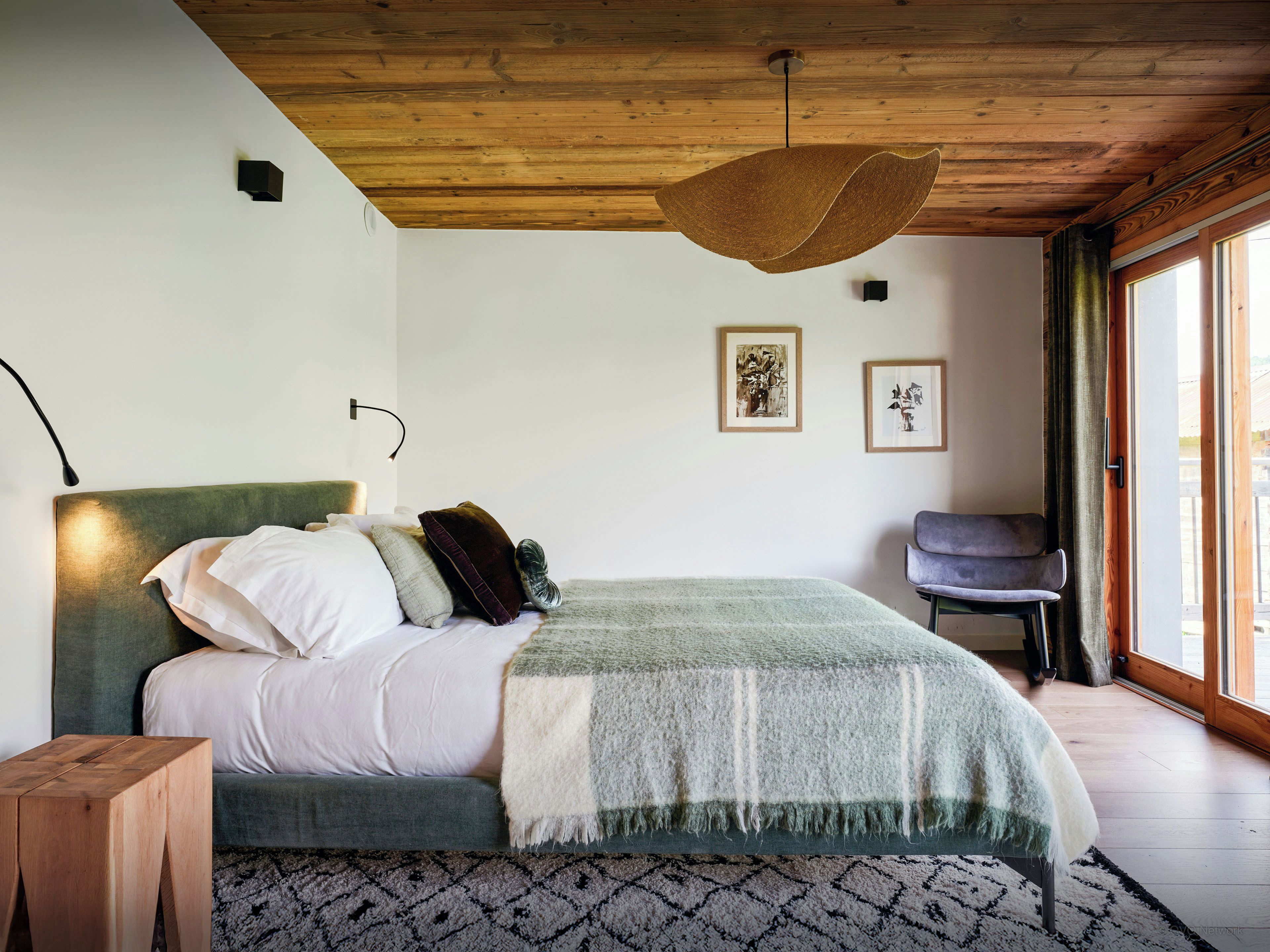 A bedroom with a bed, a wooden headboard, a hanging light fixture, a chair, and a window with curtains.