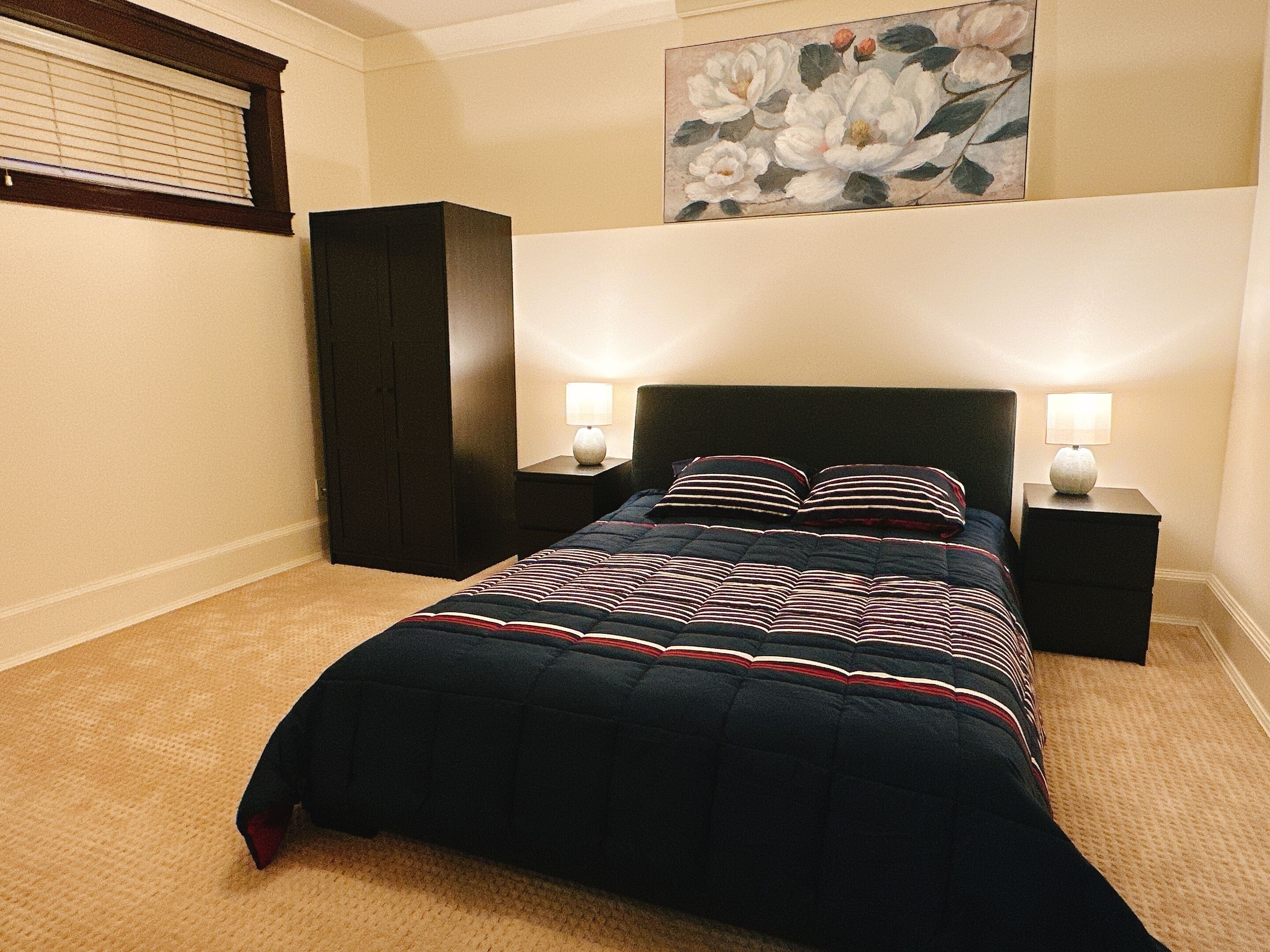 Ein Schlafzimmer mit einem dunkelbraunen Bett, zwei Nachttischlampen, einem dunklen Kleiderschrank und einem Blumenbild an der Wand.