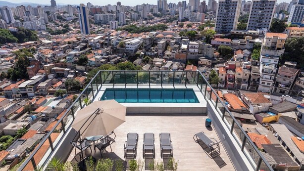 A rooftop pool with a glass balustrade, surrounded by lounge chairs and a table, overlooking a densely packed urban area with various building styles. - Spin Stays (São Paulo)