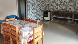 A dining area with a table covered in a floral tablecloth, surrounded by wooden chairs. A kitchen area with a stove and sink is visible in the background.