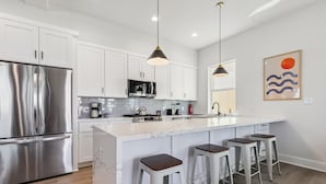 A modern kitchen with white cabinets, stainless steel appliances, a marble island with bar stools, pendant lighting, and a wall art piece.