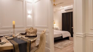 A hotel room with a dining area featuring a table set for two, a chandelier, and a decorative chair. There is a bed with a white comforter and a nightstand with a lamp. - The Versailles Suite A Designer Home in Dhaka (Dhaka)