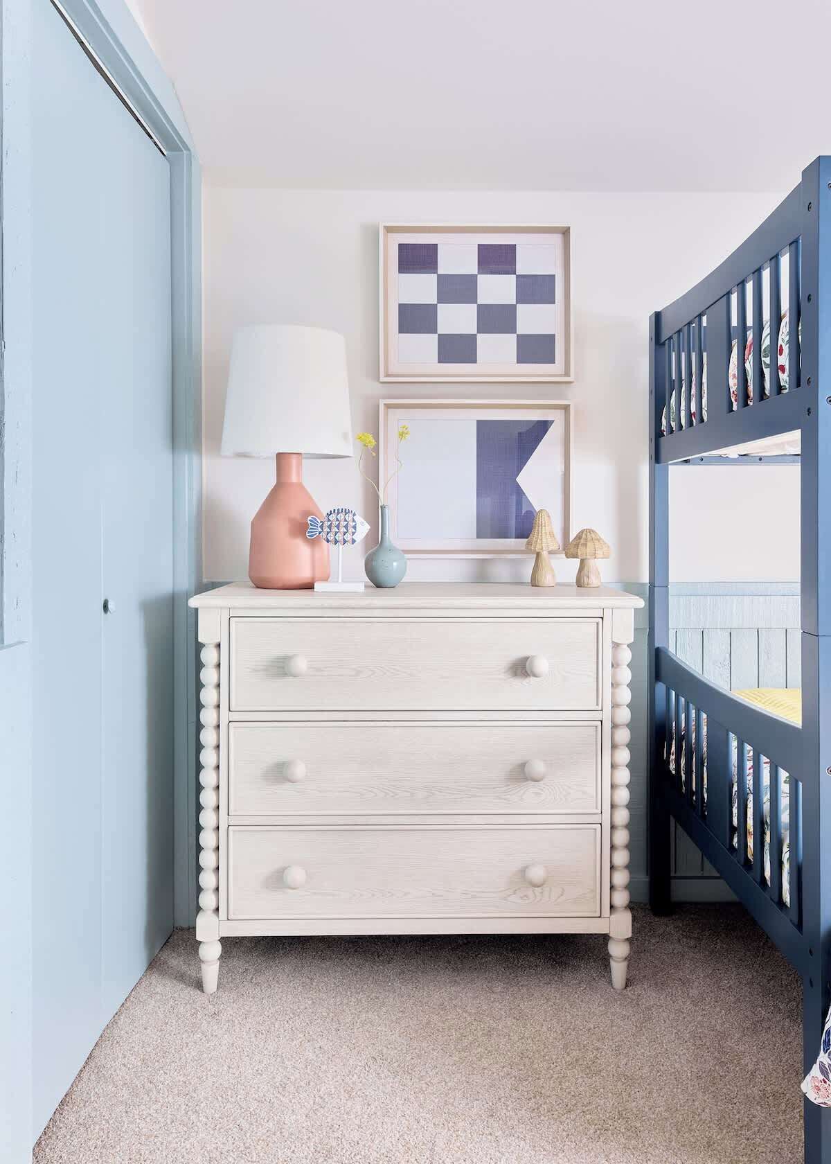 Une pièce avec une commode de couleur claire, un berceau bleu, un tableau encadré et une lampe.
