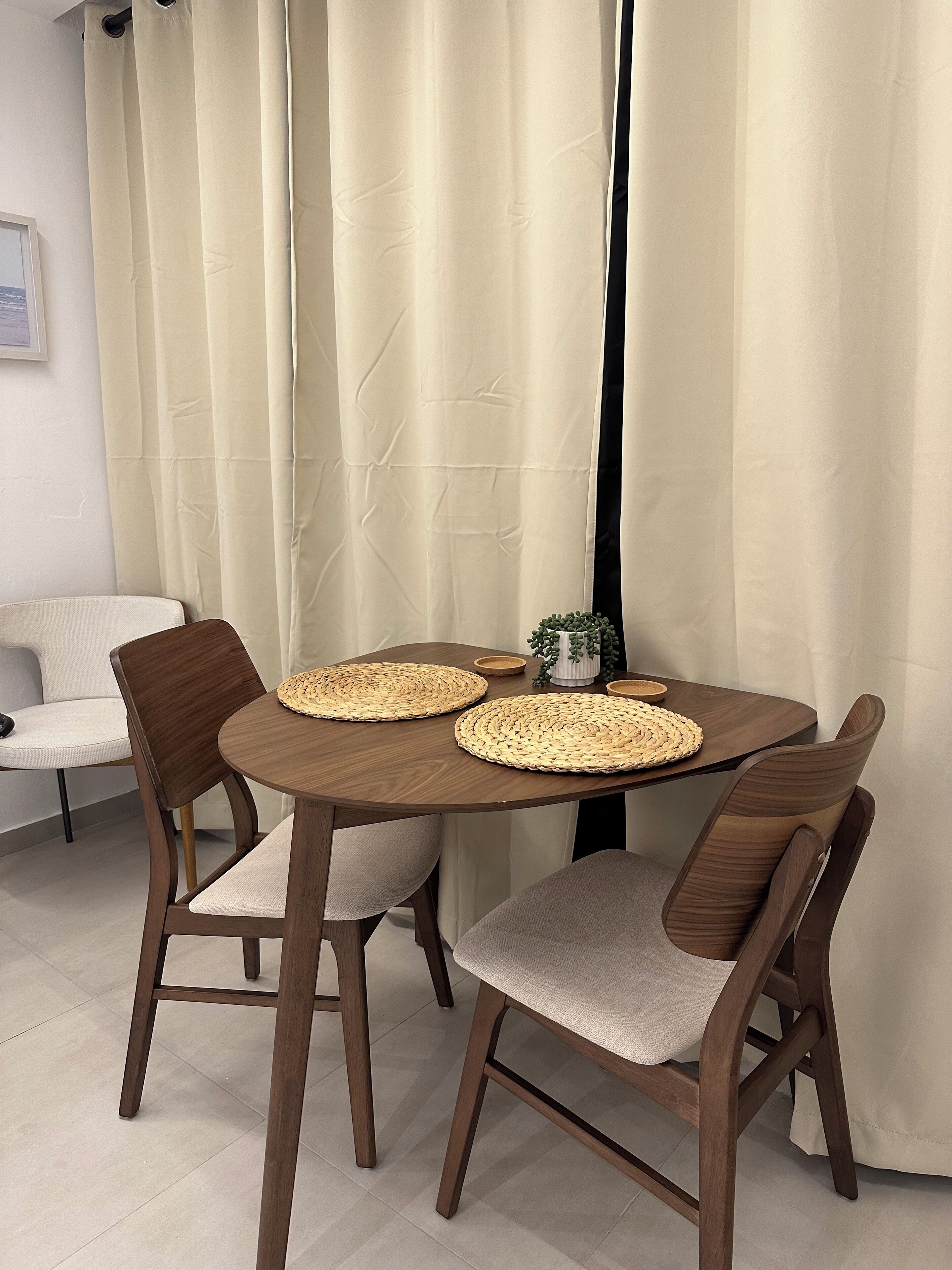Een ronde eettafel met geweven placematen, vier houten stoelen met beklede zitplaatsen, en een witte vaas met groene planten op de tafel.
