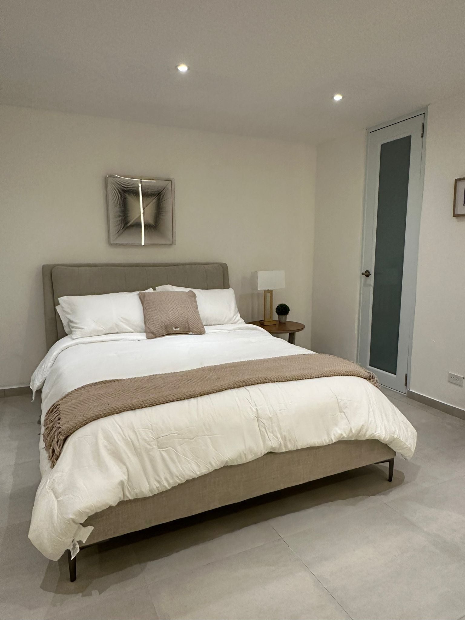 A modern bedroom with a large bed, a bedside table with a lamp, and a door with a frosted glass panel.