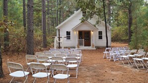 Une petite cabane blanche avec un porche, entourée d’arbres, et des chaises disposées en rangées devant.