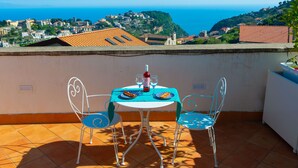 A terrace with a table and chairs, overlooking a coastal town with buildings and a view of the sea.