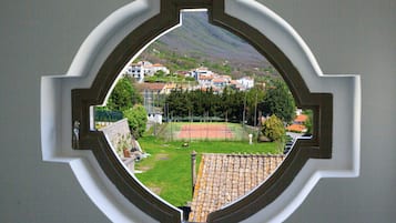 Visão pela janela com moldura decorativa, mostrando uma quadra de tênis, área verde e edifícios ao fundo.