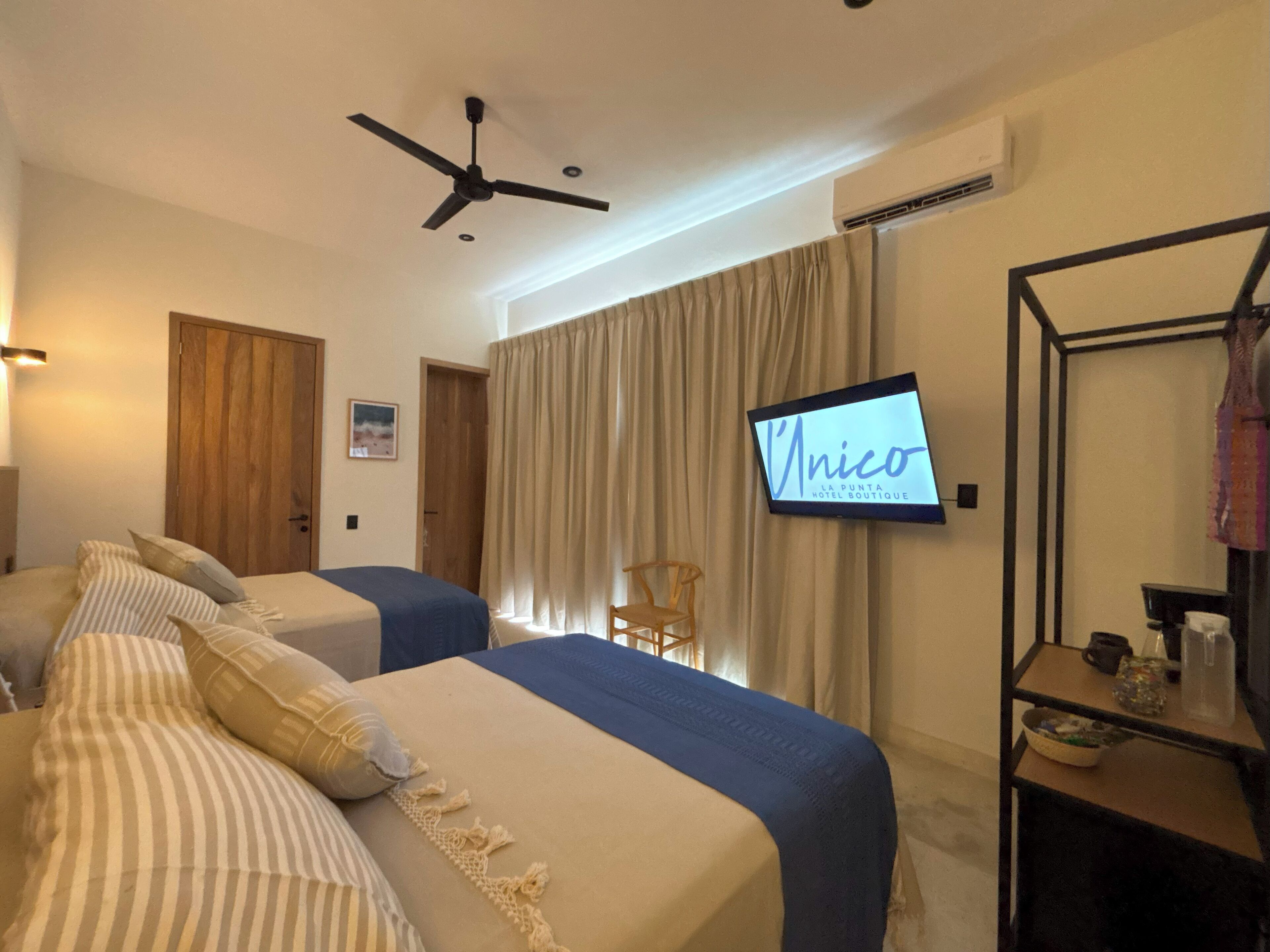 A hotel room with two beds, a ceiling fan, a TV displaying 'Unico', and a wooden door.
