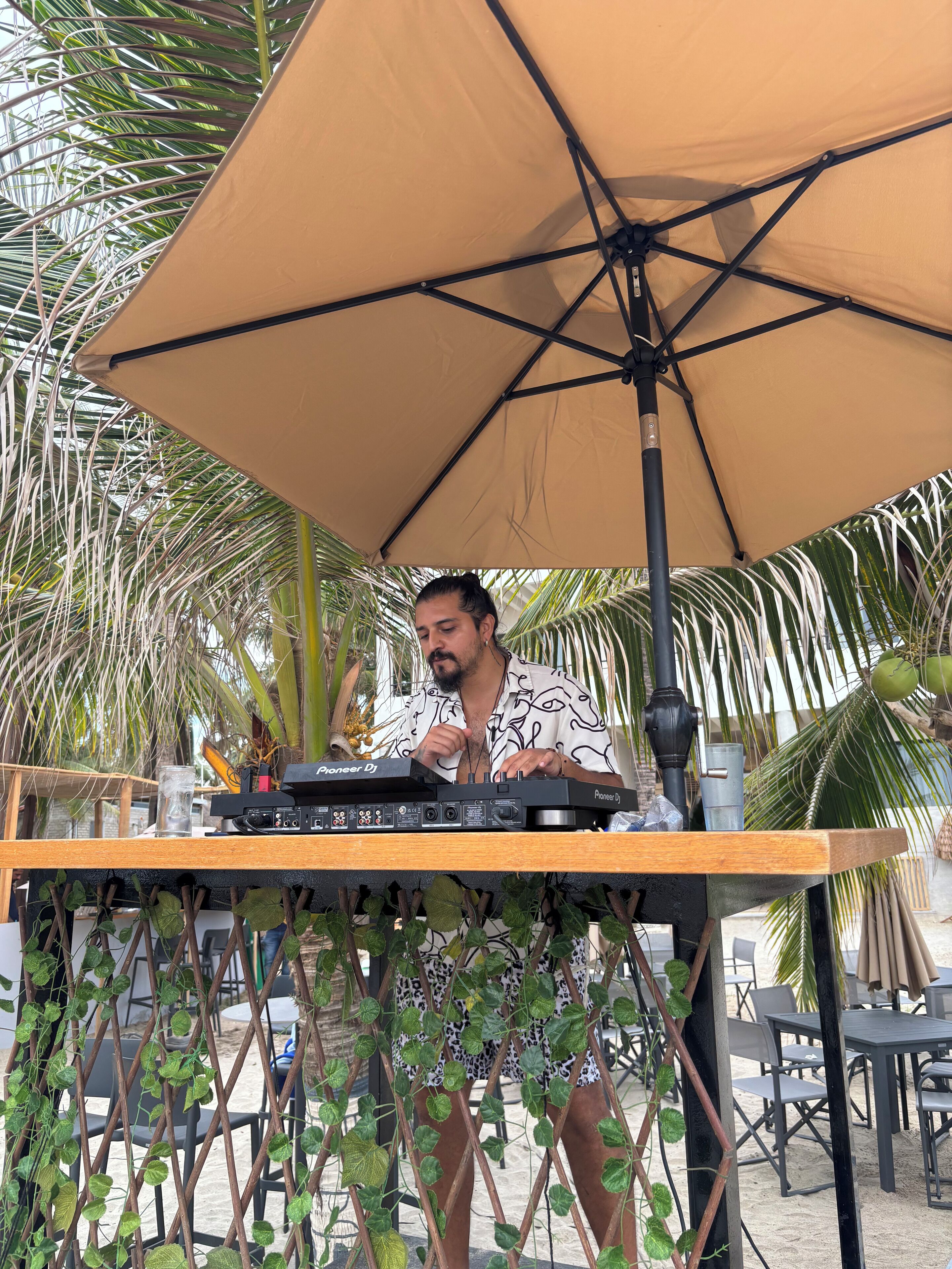Un puesto de DJ con un controlador Pioneer, un vaso y un coco sobre una mesa bajo un paraguas grande, rodeado de sillas y palmeras.