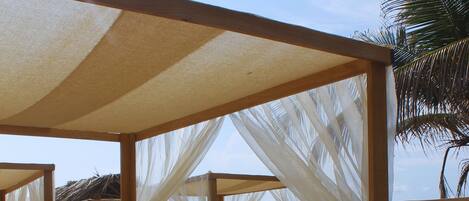 Una cabaña de playa con techo de paja, cortinas blancas y estructura de madera.