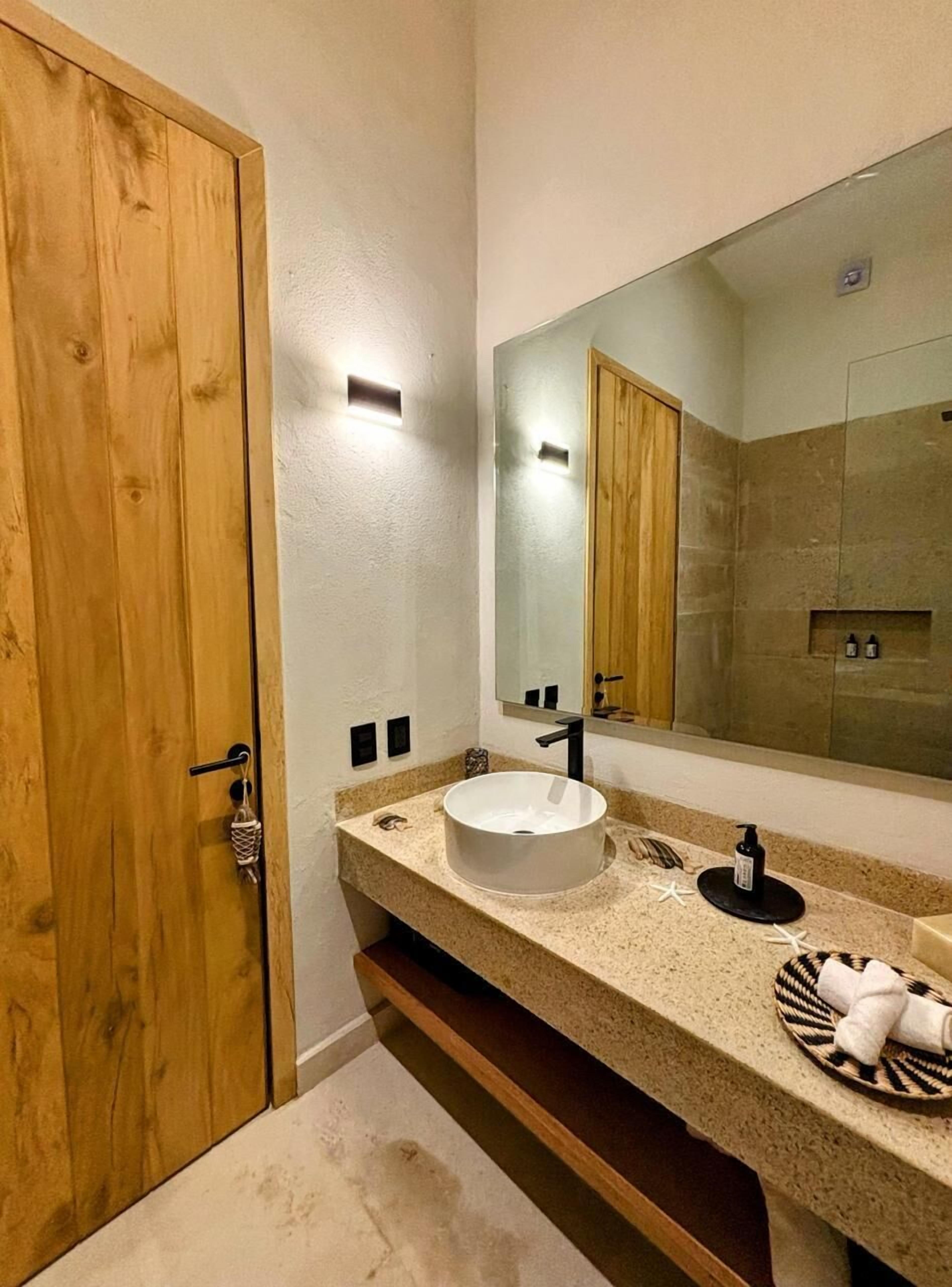 Une salle de bain avec une porte en bois, un grand miroir, un lavabo blanc et un porte-serviettes.