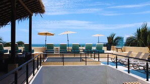 Área de piscina com espreguiçadeiras e guarda-sóis, com vista para um céu azul e o oceano.