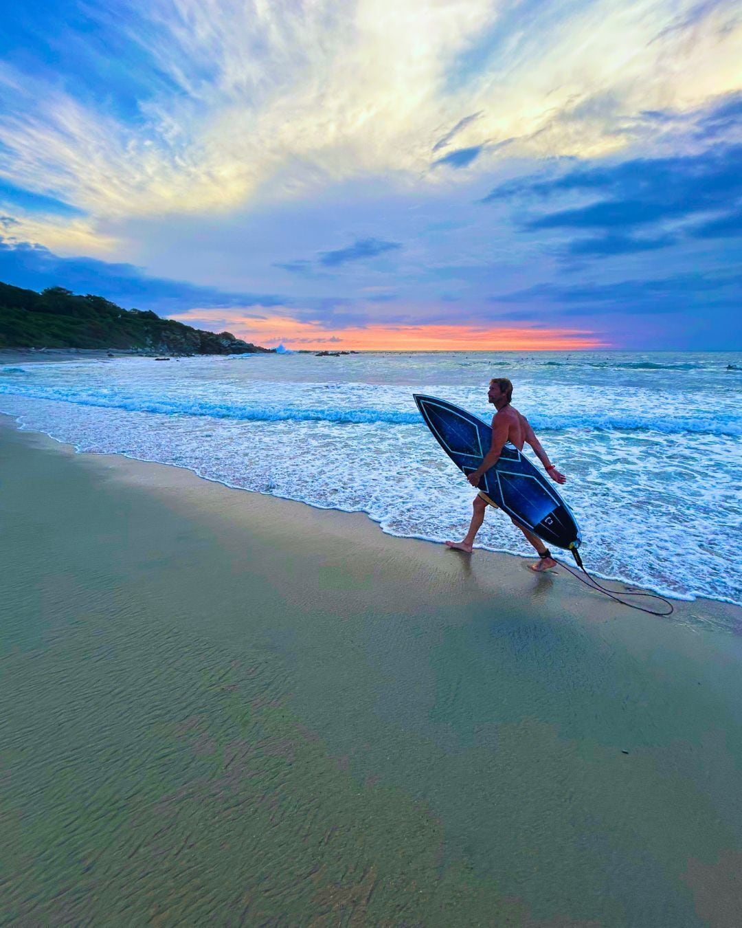 Una persona caminando en la playa con una tabla de surf, olas del océano y un cielo al atardecer.