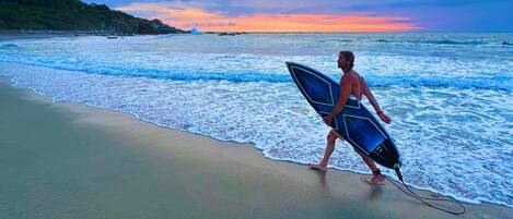 A person walking on a beach with a surfboard, ocean waves, and a sunset sky.